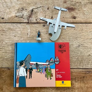 K&ouml;nnte beinhalten: Eine Anordnung von Tintin-Themen auf einer Holzoberfl&auml;che. Enthalten sind ein Buch mit dem Titel "Le Vol Air India de Tintin au Tibet", ein Modellflugzeug und eine kleine Figur. Das Buchcover zeigt einen blauen Himmel und einen roten Rand.