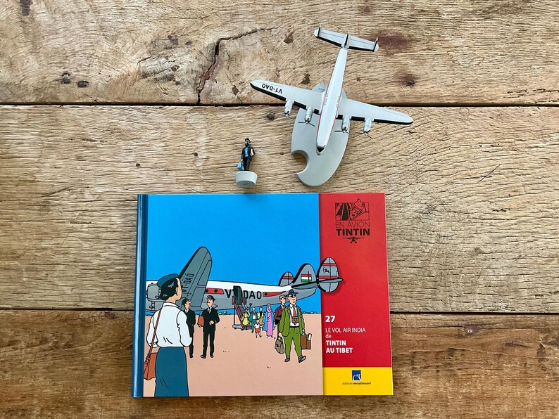 K&ouml;nnte beinhalten: Eine Anordnung von Tintin-Themen auf einer Holzoberfl&auml;che. Enthalten sind ein Buch mit dem Titel "Le Vol Air India de Tintin au Tibet", ein Modellflugzeug und eine kleine Figur. Das Buchcover zeigt einen blauen Himmel und einen roten Rand.