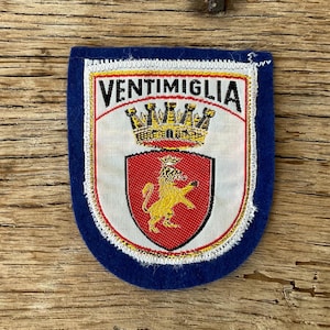 Op de afbeelding: Een blauwe patch met een wit en rood wapen met een gouden leeuw en kroon. Het woord "VENTIMIGLIA" staat in zwarte letters boven het wapen gedrukt. De patch heeft een witte rand.