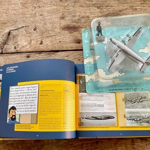 K&ouml;nnte beinhalten: Ein offenes Buch mit farbigen Seiten und Illustrationen, neben einem Spielzeugflugzeug in einer Plastikh&uuml;lle. Das Buch hat Text und Bilder, und das Flugzeug ist wei&szlig; und silberfarben mit dem Text "VT-BAD".