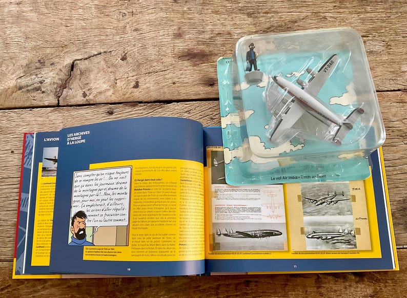 K&ouml;nnte beinhalten: Ein offenes Buch mit farbigen Seiten und Illustrationen, neben einem Spielzeugflugzeug in einer Plastikh&uuml;lle. Das Buch hat Text und Bilder, und das Flugzeug ist wei&szlig; und silberfarben mit dem Text "VT-BAD".
