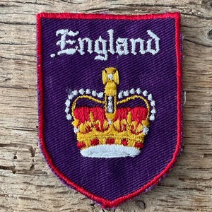 May include: A shield-shaped patch in purple with a red border. It features the word "England" in white script above a detailed crown in gold, red, and white. A decorative emblem.