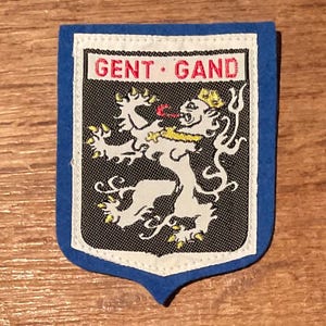 May include: A blue and white patch with the text "GENT · GAND" in red. The patch features a white lion with a yellow crown and accents on a black background.