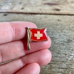 Könnte beinhalten: Ein kleiner, rot-weißer Schweizer Flaggen-Anstecker mit goldfarbenem Mast. Die Flagge zeigt ein weißes Kreuz auf rotem Grund. Der Anstecker ist wahrscheinlich aus Metall und hat eine glänzende Oberfläche. Der Anstecker ist eine Miniaturdarstellung der Schweizer Flagge.