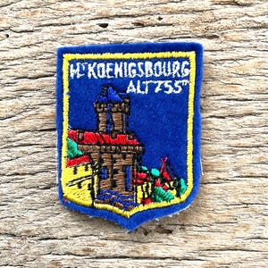 May include: A blue embroidered patch featuring a castle tower with a red roof and yellow accents. The text reads "H KOENIGSBOURG ALT 755". The patch has a yellow border and a shield-like shape, showcasing detailed stitching of the stone structure.