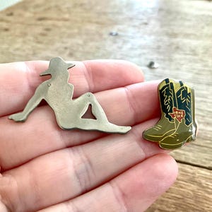 Op de afbeelding: Twee metalen spelden: een in de vorm van een liggende figuur met een hoed en de andere een paar cowboylaarzen met een Lone Star-ontwerp. De laarzen zijn olijfgroen met zwarte en gouden accenten.