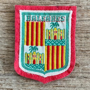 Op de afbeelding: Een rechthoekige geborduurde patch met een rode vilten rand met het wapen van de Balearen. Het ontwerp bevat vier verticale rode strepen op een gele achtergrond en een kasteel met een palmboom, met de tekst BALEARES.