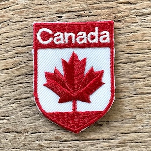 May include: Embroidered patch with the word "Canada" in white on a red and white shield design. The patch features a red maple leaf, a symbol of Canada, and is edged in red.