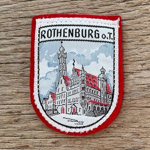 Könnte beinhalten: Rechteckiger Aufnäher mit rotem Rand und detaillierter Illustration eines Gebäudes mit roten Dächern und einem hohen Turm. Der Aufnäher trägt den Text "ROTHENBURG O.T." in schwarzen Buchstaben.