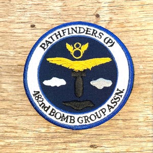 Op de afbeelding: Een cirkelvormige patch met de tekst "PATHFINDERS (P)" en "482nd BOMB GROUP ASSN." in witte letters op een marineblauwe achtergrond. De patch heeft een geel vleugelontwerp, een zwarte bom en twee witte wolkvormen. De rand is wit en blauw.