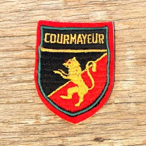 Könnte beinhalten: Gestickter Aufnäher mit dem Wort "COURMAYEUR" in goldener Schrift. Der Aufnäher zeigt ein Schilddesign mit schwarzem Oberteil, rotem Unterteil und einem goldenen Löwen. Der Aufnäher ist grün umrandet.