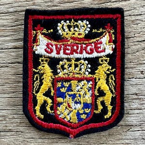 Op de afbeelding: Een rechthoekige geborduurde patch met een zwarte achtergrond en een rode rand. Het toont een centraal schild met blauwe en gele details, geflankeerd door twee gele leeuwen, bekroond met een kroon en een witte banner met de tekst SVERIGE.