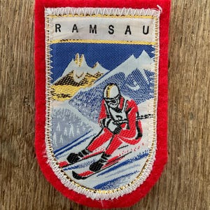 Könnte beinhalten: Vintage Ramsau-Aufnäher mit rotem Rand und Skifahrer-Illustration. Der Aufnäher zeigt das Wort "RAMSAU" oben, mit einer blau-weißen Berglandschaft und einem Skifahrer in rotem Anzug.