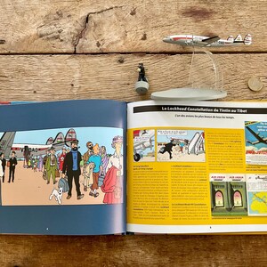 K&ouml;nnte beinhalten: Ein offenes Buch mit einem Comic-Strip, der Menschen und ein Flugzeug zeigt. Ein Modellflugzeug und eine Figur liegen auf einer Holzoberfl&auml;che. Der Titel des Buches lautet "L'Avion".