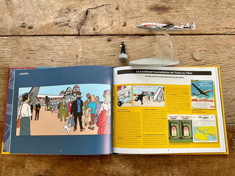 K&ouml;nnte beinhalten: Ein offenes Buch mit einem Comic-Strip, der Menschen und ein Flugzeug zeigt. Ein Modellflugzeug und eine Figur liegen auf einer Holzoberfl&auml;che. Der Titel des Buches lautet "L'Avion".