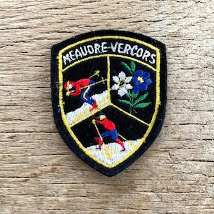以下が含まれることがあります： 黒いシールド型の刺繍パッチで、白で「MEAUDRE-VERCORS」の文字が描かれています。 パッチは黄色の縁取りがあり、3つのセクションにスキーヤーと花が描かれています。 スキーヤーは赤と青の衣装を着ています。