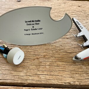 K&ouml;nnte beinhalten: Ein Modellflugzeug mit dem Text "VT-DAO" und eine Figur einer Figur aus der Tintin-Serie. Eine graue ovale Basis mit franz&ouml;sischem Text, darunter "Le vol Air India Tintin au Tibet" und "Page 6 - &Eacute;chelle 1:250".