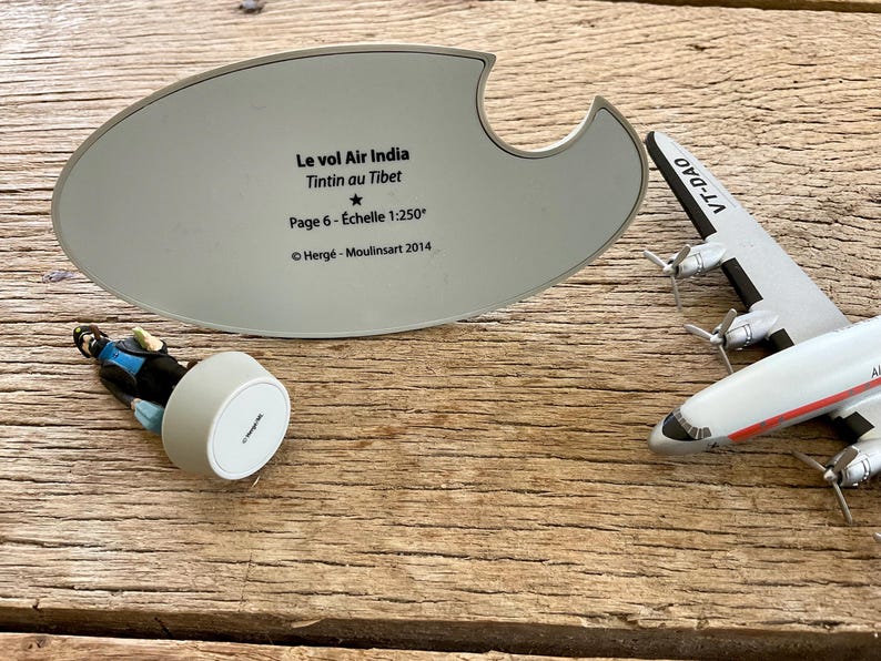 K&ouml;nnte beinhalten: Ein Modellflugzeug mit dem Text "VT-DAO" und eine Figur einer Figur aus der Tintin-Serie. Eine graue ovale Basis mit franz&ouml;sischem Text, darunter "Le vol Air India Tintin au Tibet" und "Page 6 - &Eacute;chelle 1:250".