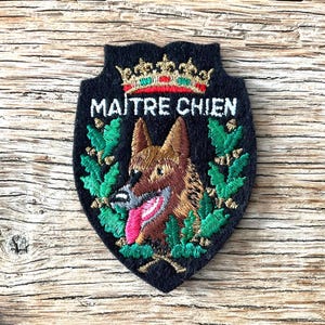 May include: Black shield-shaped patch with the text "MAITRE CHIEN" in white. The patch features a German Shepherd dog illustration, a gold crown, and green leaf accents. The dog has a pink tongue.