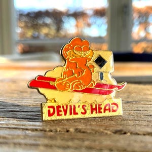 May include: A vintage pin featuring an orange cartoon cat character on a red and white cloud, with a gold border. The pin reads "DEVIL'S HEAD" in red letters.