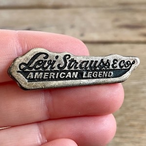 Może przedstawiać: Vintage metalowa przypinka Levi Strauss & Co. z napisami "Levi Strauss & Co." i "AMERICAN LEGEND" w czarnych literach na srebrnym tle. Przypinka jest trzymana w dłoni.