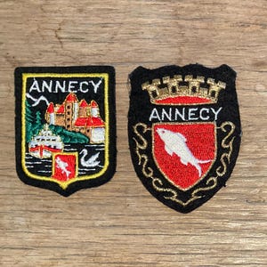 Può includere: Due toppe ricamate con la scritta "ANNECY". Una raffigura un castello e un lago, l'altra un pesce su uno scudo rosso. Entrambe le toppe hanno un bordo nero e dettagli dorati.