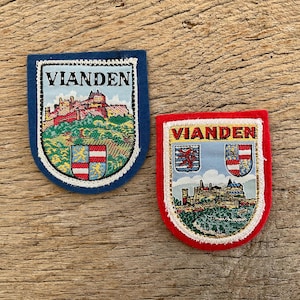 Op de afbeelding: Twee geborduurde patches met het woord "Vianden". Eén is blauw, met een kasteel en wapenschild. De andere is rood, met een vergelijkbare kasteelillustratie en wapenschild.