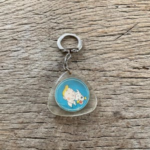 Pu&ograve; includere: Un portachiavi triangolare trasparente con un'immagine circolare di un personaggio dei cartoni animati con i capelli biondi e un cane bianco su sfondo blu. Il portachiavi ha un anello e una catena color argento.