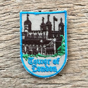 Op de afbeelding: Geborduurde patch met de Tower of London in bruin en groen tegen een lichtgrijze achtergrond. De patch heeft een blauwe rand en de woorden "Tower of London" in blauw.