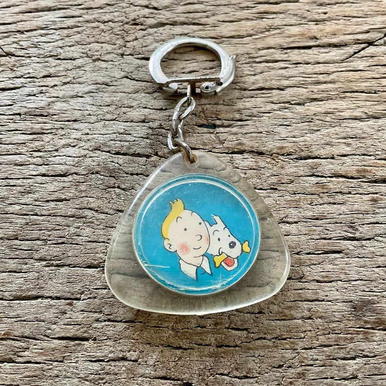 Pu&ograve; includere: Un portachiavi vintage con un'immagine a cartoni animati di un ragazzo con i capelli gialli e un cane bianco su sfondo blu. Il portachiavi ha una base in plastica trasparente triangolare e un portachiavi in metallo argentato.