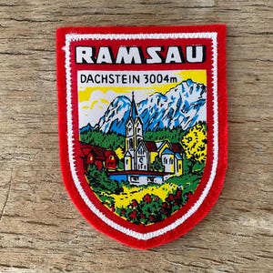 Könnte beinhalten: Ein roter, schildförmiger Aufnäher mit weißem Rand und dem Wort "RAMSAU" oben. Darunter steht "DACHSTEIN 3004m". Der Aufnäher zeigt eine farbenfrohe Illustration einer Kirche, Häusern, Bergen und Bäumen.