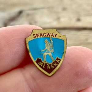 May include: A vintage Skagway Alaska souvenir pin. The shield-shaped pin features a blue center with a gold figure and the words "SKAGWAY" and "ALASKA". The pin is held in a person's hand.
