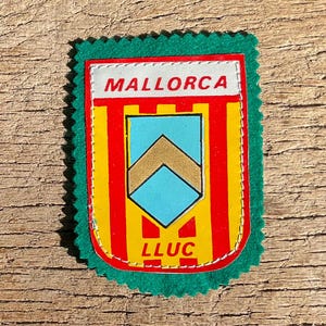 Op de afbeelding: Een rechthoekige patch met een groene geschulpte rand. De patch heeft een ontwerp met rode en gele verticale strepen, een lichtblauwe ruitvorm en de woorden "MALLORCA" bovenaan en "LLUC" onderaan.