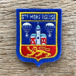 May include: A blue, shield-shaped patch with gold trim. The patch features a church, parachutes, and text that reads "ST MERE EGLISE". A red and gold emblem is at the bottom.