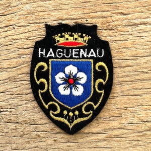 Op de afbeelding: Een zwart en goud geborduurde patch met het woord "HAGUENAU" bovenaan. De patch heeft een blauw schild met een witte bloem en een rood centrum. De patch is gevormd als een schild.