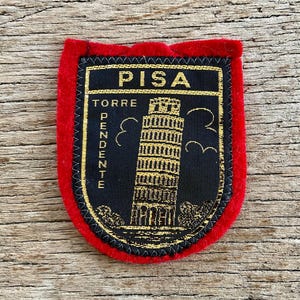 Op de afbeelding: Een schildvormige geborduurde patch met een felrode vilten rand. Het midden heeft een zwarte achtergrond met gouden metaaldraad die de Toren van Pisa afbeeldt, met de tekst PISA en TORRE PENDENTE in goud geborduurd.