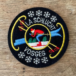 Op de afbeelding: Een ronde zwarte geborduurde patch met een skiër in een rode jas en blauwe broek, omringd door een gele ring. De tekst luidt LA SCHLUCHT VOSGES, geaccentueerd door witte sneeuwvlokmotieven en gele ski-uitrusting op een donkere achtergrond.