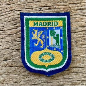 May include: A shield-shaped embroidered patch featuring the text MADRID in green on a yellow banner. The design includes a yellow dragon on a blue background, a green bear standing by a tree on a white background, and a green wreath on yellow.