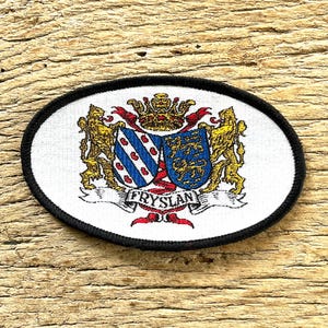 Op de afbeelding: Ovale patch met zwarte rand met een heraldisch ontwerp. Het ontwerp omvat een schild met blauwe en rode elementen, een kroon en het woord "FRYSLAN" op een spandoek. Twee gouden leeuwen flankeren het schild.