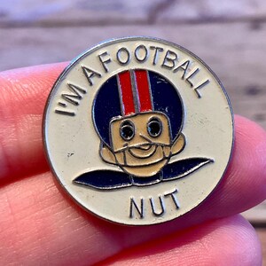 Op de afbeelding: Een ronde, witte pin met de tekst "I'M A FOOTBALL NUT". De pin toont een cartoon American footballspeler met een blauwe helm met rode strepen. De speler heeft een bruin gezicht en een lachende uitdrukking.
