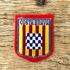 Op de afbeelding: Een rood, schildvormig embleem met de tekst "COSTA BRAVA" in blauw en wit. De patch heeft verticale gele en rode strepen en een centraal vierkant met een blauw-wit geblokt patroon.