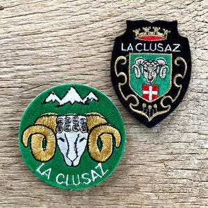 Op de afbeelding: Twee geborduurde patches met de tekst "LA CLUSAZ". Eén is rond en groen met een rammenkop en bergontwerp. De andere is schildvormig, zwart met gouden rand en heeft een rammenkop en een rood kruis.