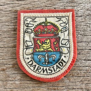 Puede incluir: Un parche de Darmstadt con borde rojo e interior crema. El parche presenta un escudo de armas con una corona, un león y una flor de lis, con la palabra "DARMSTADT" en semicírculo.
