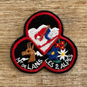 Op de afbeelding: Geborduurde patch met een zwarte rand en drie met elkaar verbonden secties. De patch toont een skiër, een lama en bloemelementen, met de tekst "MT DE LANS" en "LES 2 ALPES".