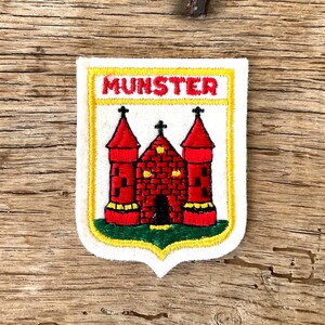 Op de afbeelding: Geborduurde patch met een rood kasteel met twee torens en een centraal gebouw, tegen een witte achtergrond. Het woord "MUNSTER" staat in gele letters bovenaan. De patch heeft een gele rand en een groene basis.