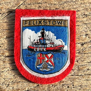 May include: A rectangular patch with a red border and a detailed embroidered design. The design features a ship, lighthouse, and the text "FELIXSTOWE" and "CORK". The patch has a white border around the central design.