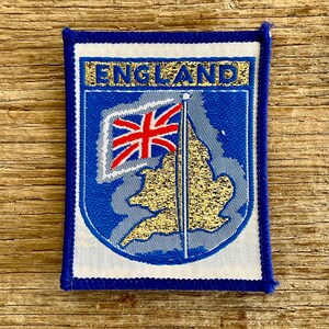 Op de afbeelding: Een rechthoekige geborduurde patch met een blauwe rand en een witte achtergrond. Het toont het woord ENGLAND in gouden letters boven een blauw schild met een gouden kaart van Engeland, een vlaggenmast en een rood-wit-blauwe Union Jack vlag.