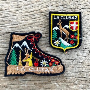 Op de afbeelding: Twee geborduurde patches met de tekst "LA CLUSAZ". Eén patch heeft de vorm van een wandelschoen met berg-, boom- en bloemdetails. De andere is een schild met een hert, bergen en een Zwitserse vlag.