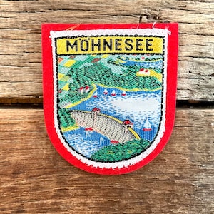以下が含まれることがあります： 上部に「MOHNESEE」の文字が入った赤、白、黄色のパッチ。パッチには、橋、湖、ヨットのあるカラフルな刺繍風景が描かれています。パッチは木製の表面にあります。