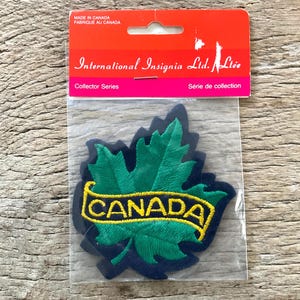 Op de afbeelding: Een groen geborduurd esdoornblad-patch met een gele "CANADA"-banner op een marineblauwe achtergrond. De verpakking toont "International Insignia Ltd. Ltée" en "Collector Series". Gemaakt in Canada.
